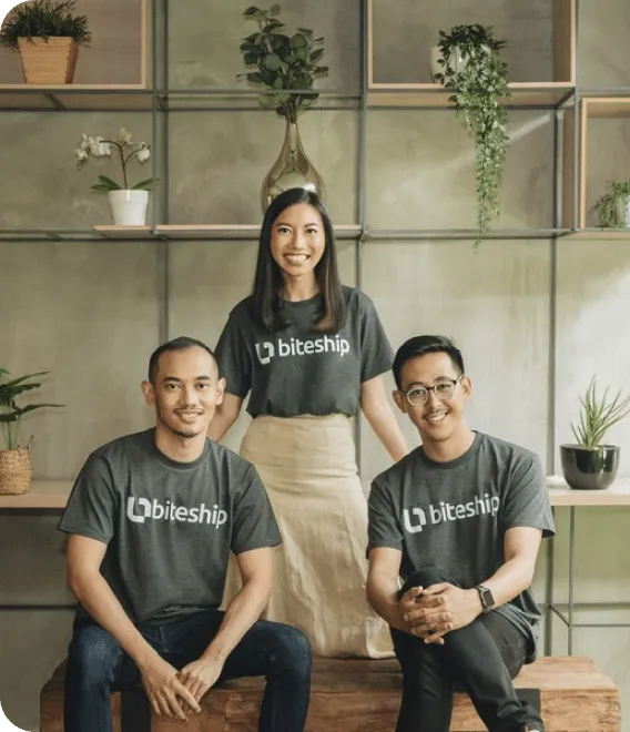 Tiga orang berpakaian kaos "biteship" tersenyum sambil duduk dan berdiri berjejer, dikelilingi rak berisi tanaman hias di interior kantor bernuansa hangat.